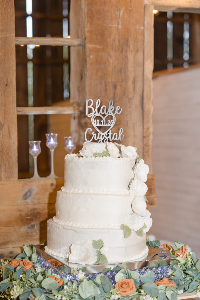 A white wedding cake with a silver "Blake and Crystal cake topper from their Barn at Whispering Pines wedding