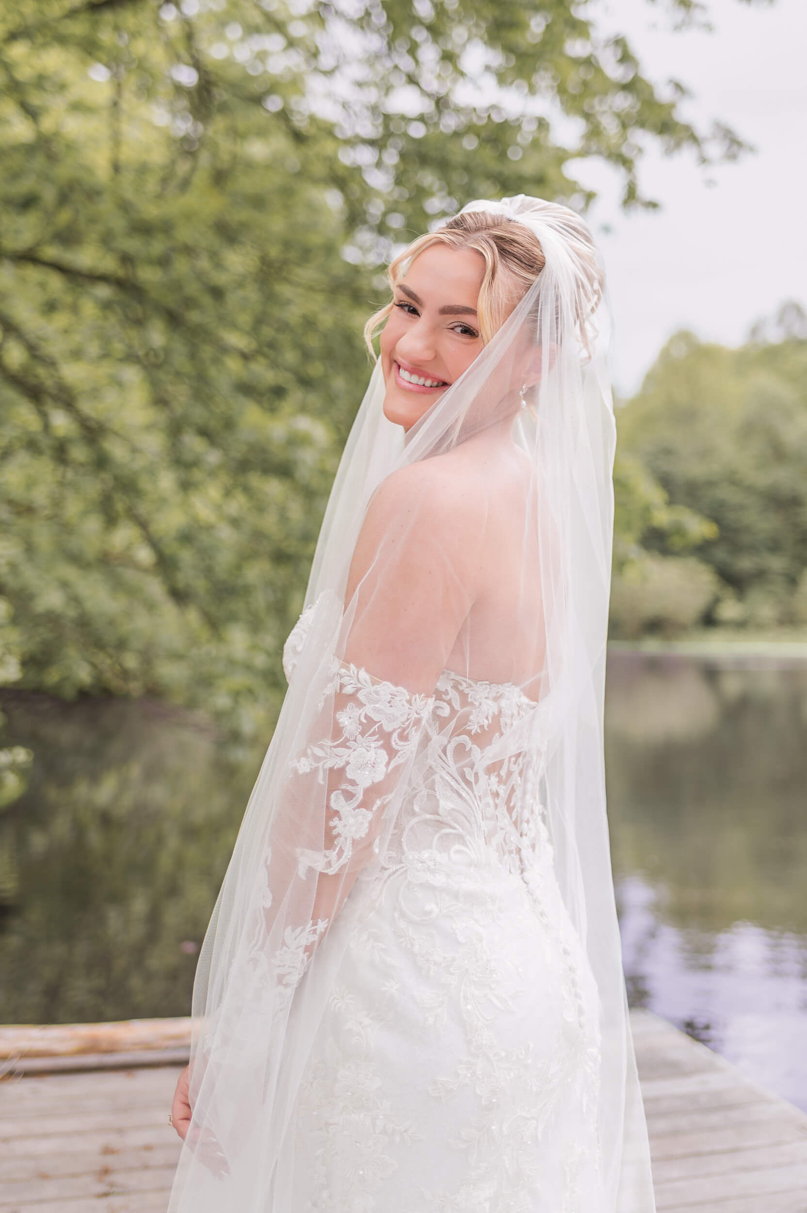 A happy bride smiles over her shoulder in her veil and lace gown on a dock at one of the outdoor wedding venues in PA