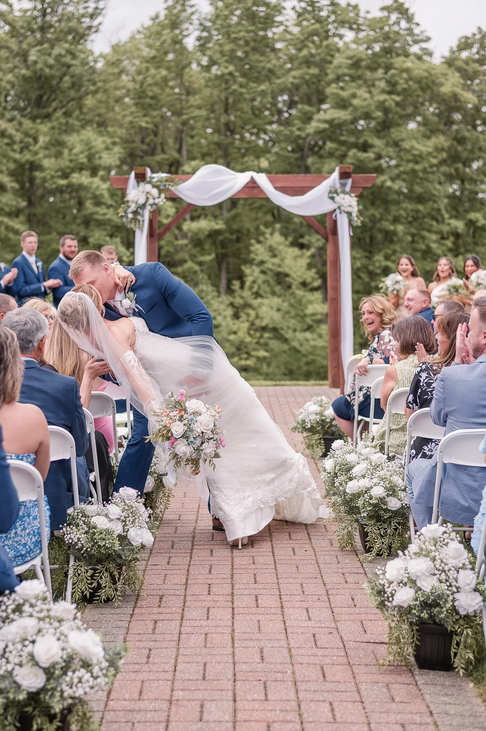 A groom in a blue suit dips and kisses his bride in the brick aisle as guests clap at one of the outdoor wedding venues in PA