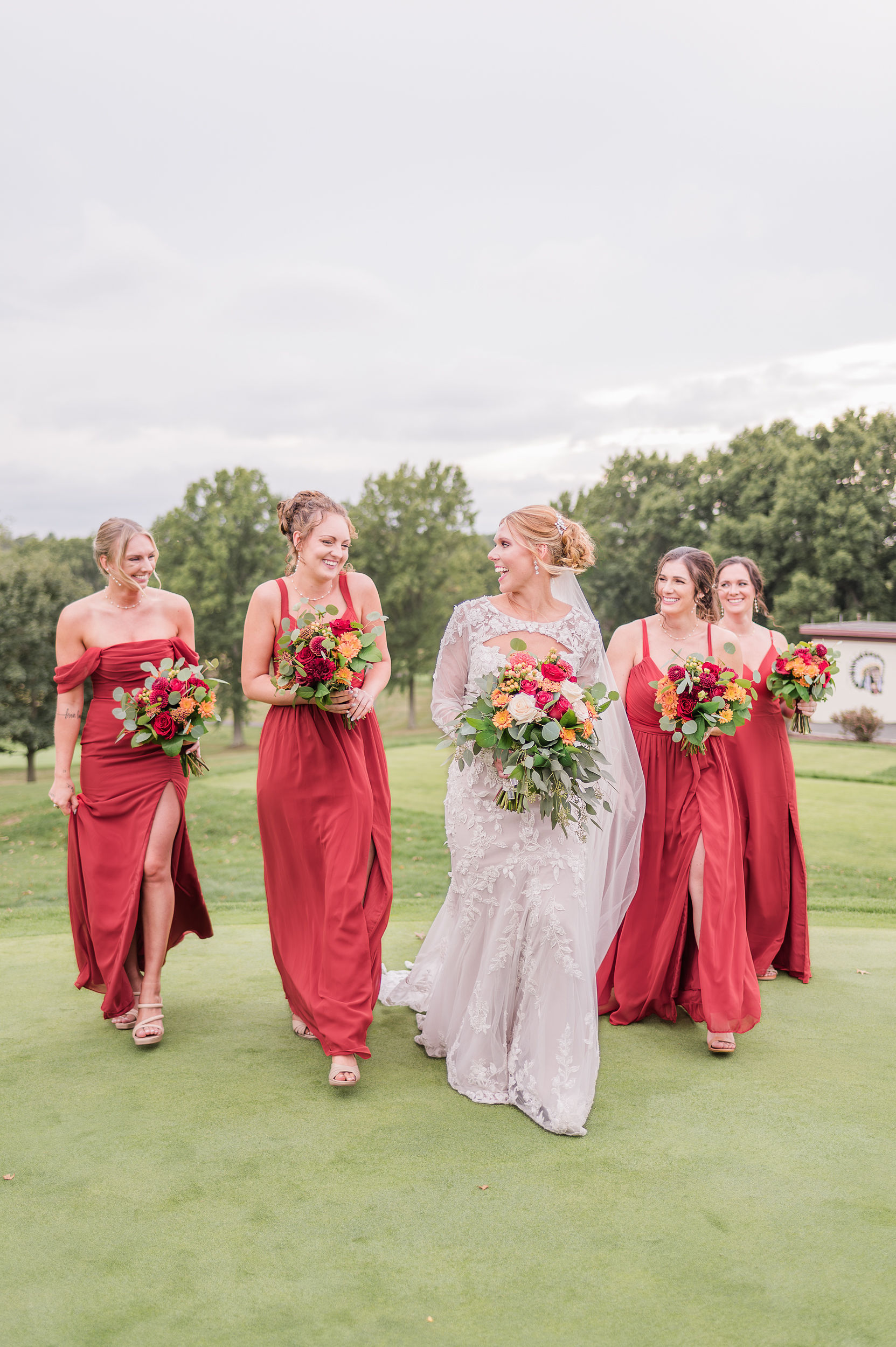 A bride laughs with a bridesmaid while walking them across a golf green in red dresses