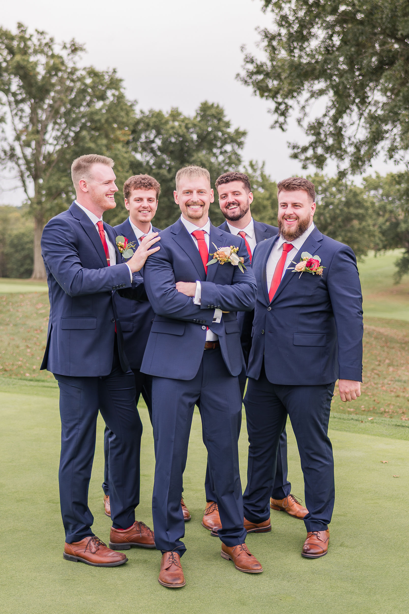 A groom smiles big while standing in his blue suit and red tie with his matching groomsmen