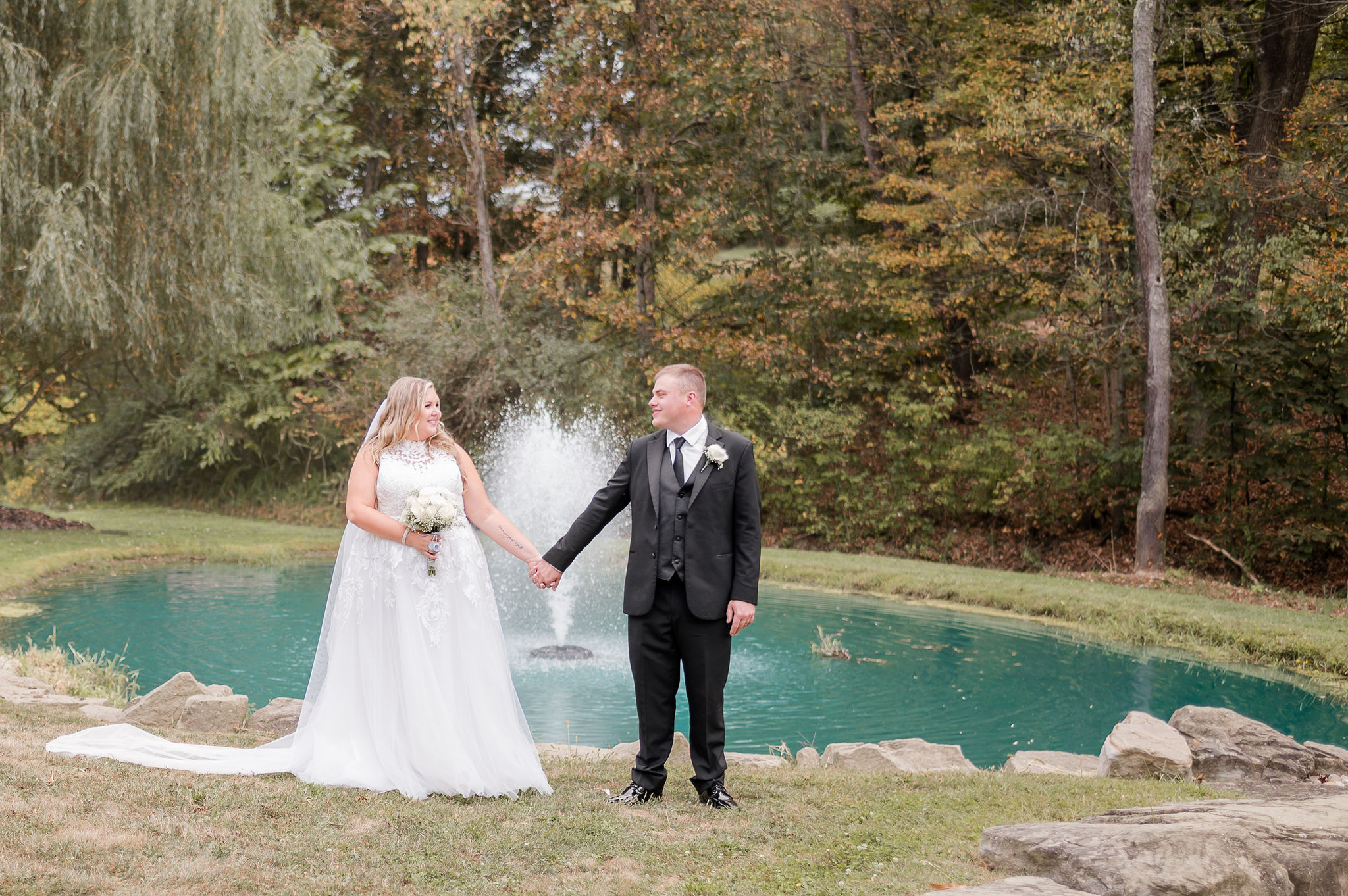 A bride and groom hold hands by a pond and willow tree smiling at each other at one of the outdoor wedding venues in PA