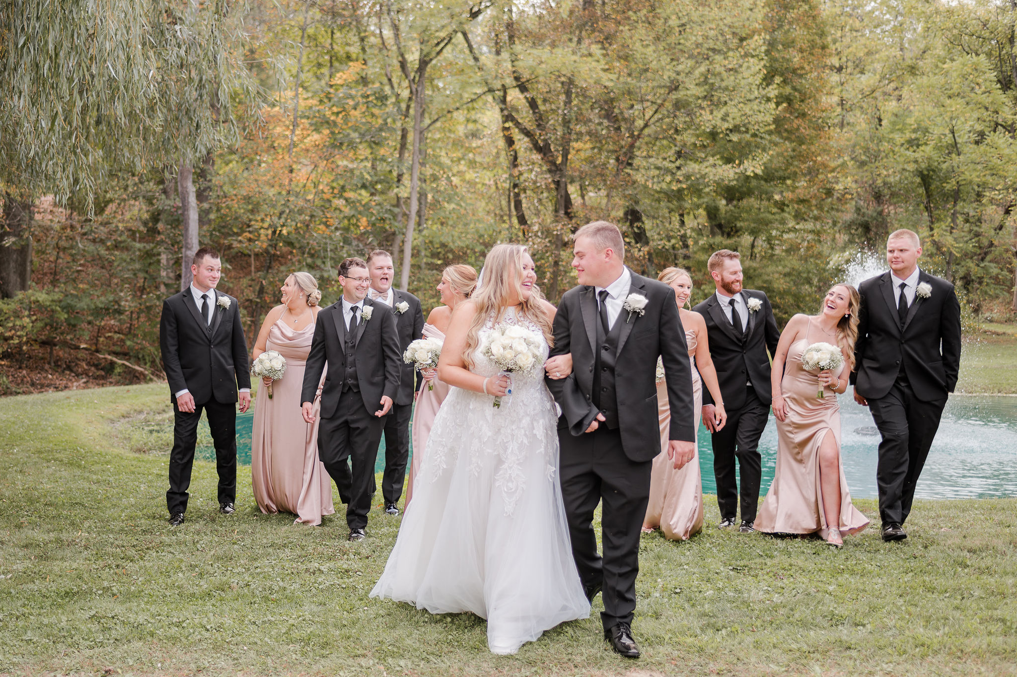 Newlyweds walk in the lawn by a pond with their wedding party wearing black and pink