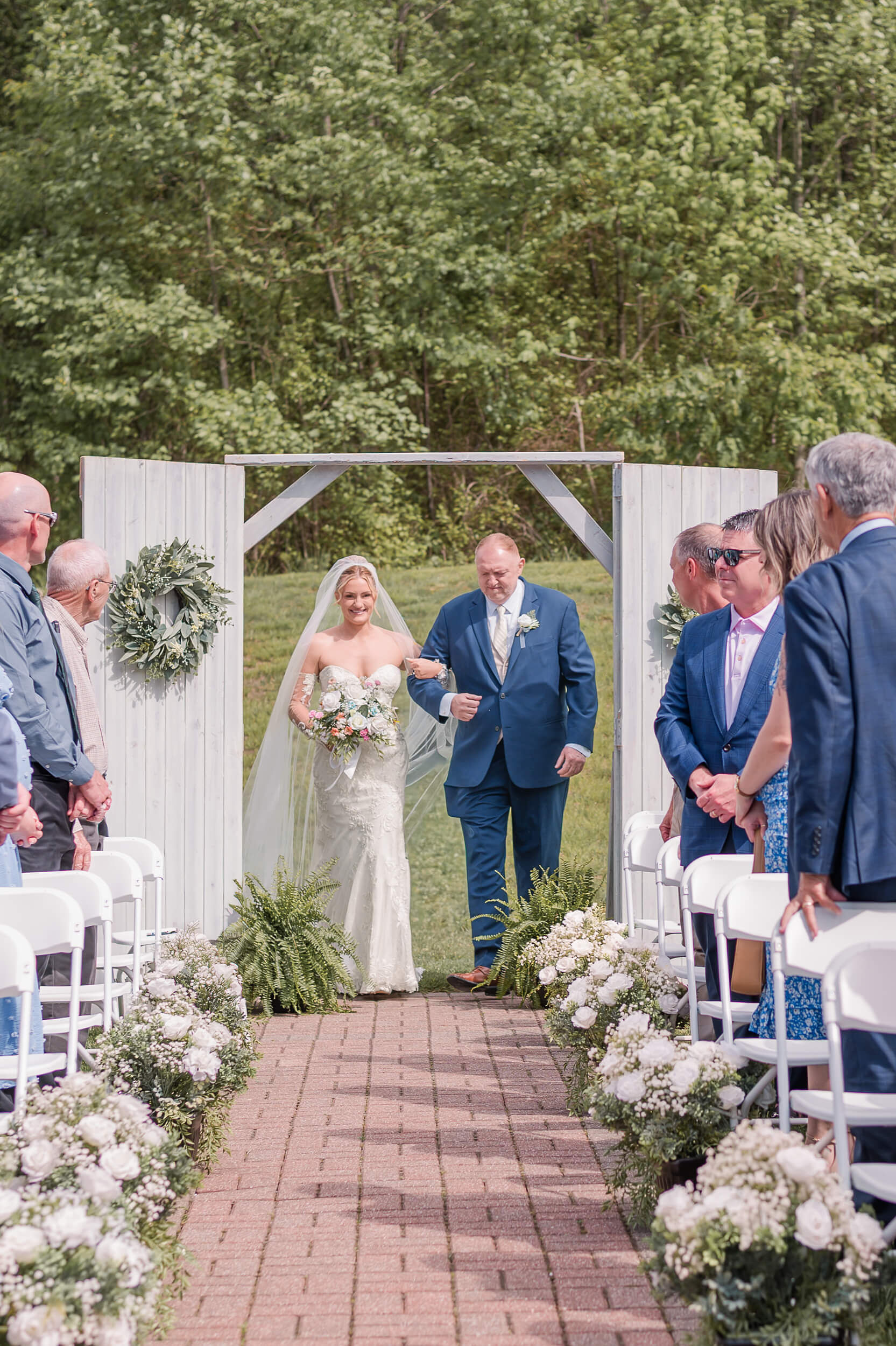 A bride walks down the aisle at one of the outdoor wedding venues in PA with her father in a blue siit as guests turn to look