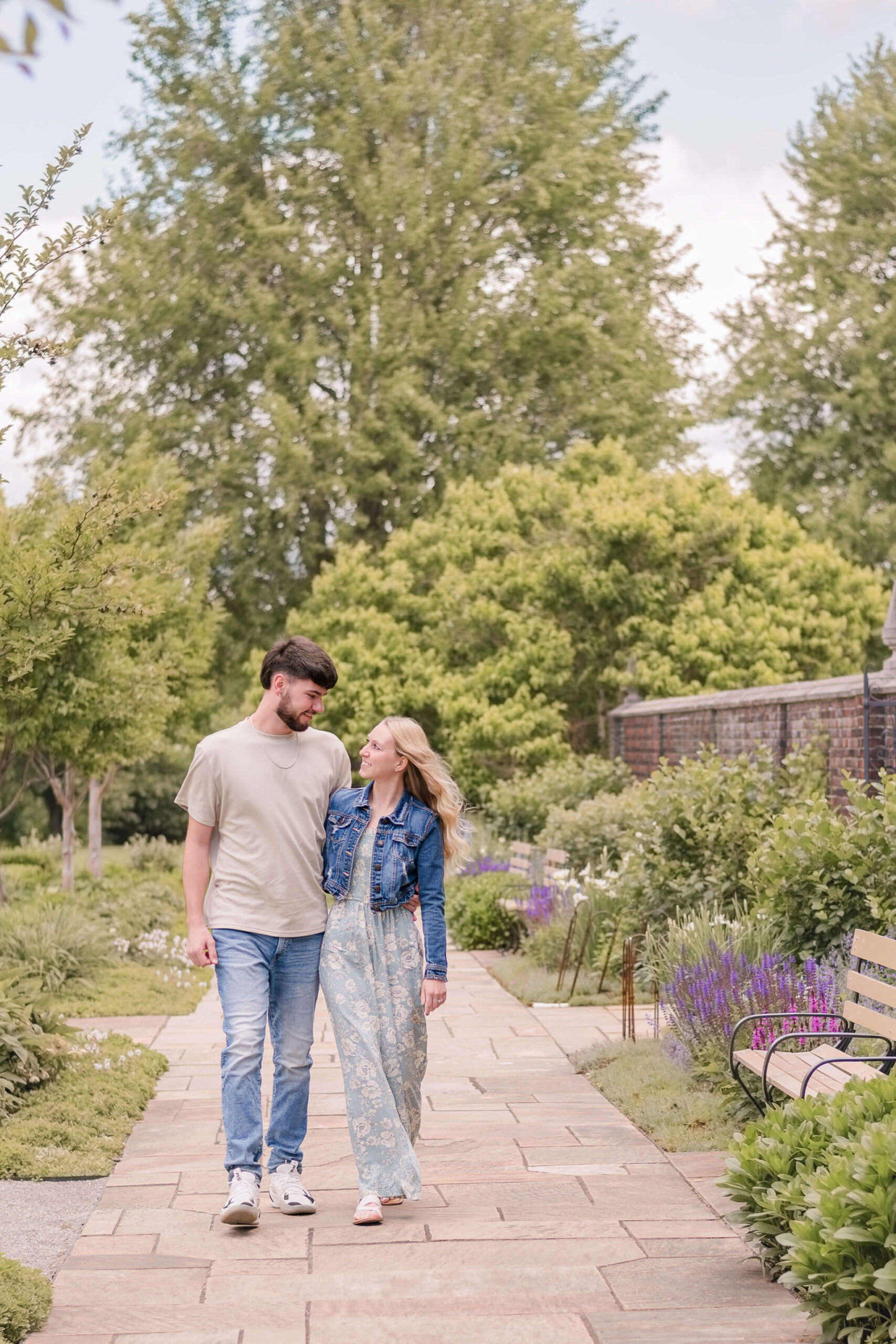 A couple walks while smiling at each other in jeans and a blue dress through a lively flower garden during their Pittsburgh engagement photos