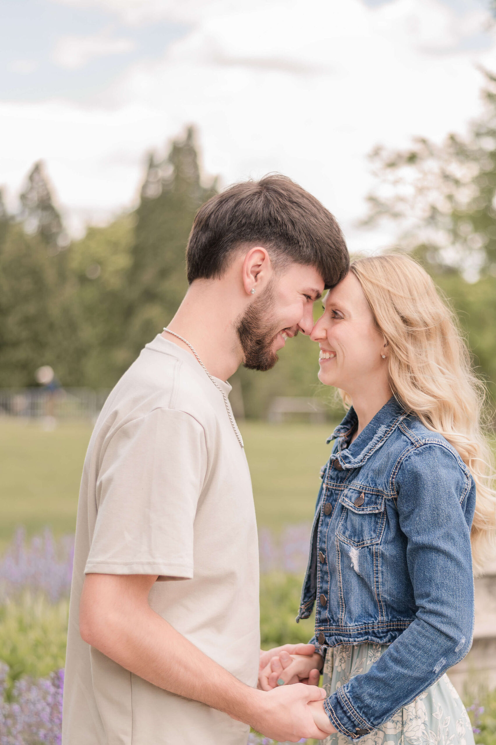 A newly engaged couple hold hands and touch foreheads in a tan shirt and denim jacket over dress in a garden during Pittsburgh engagement photos