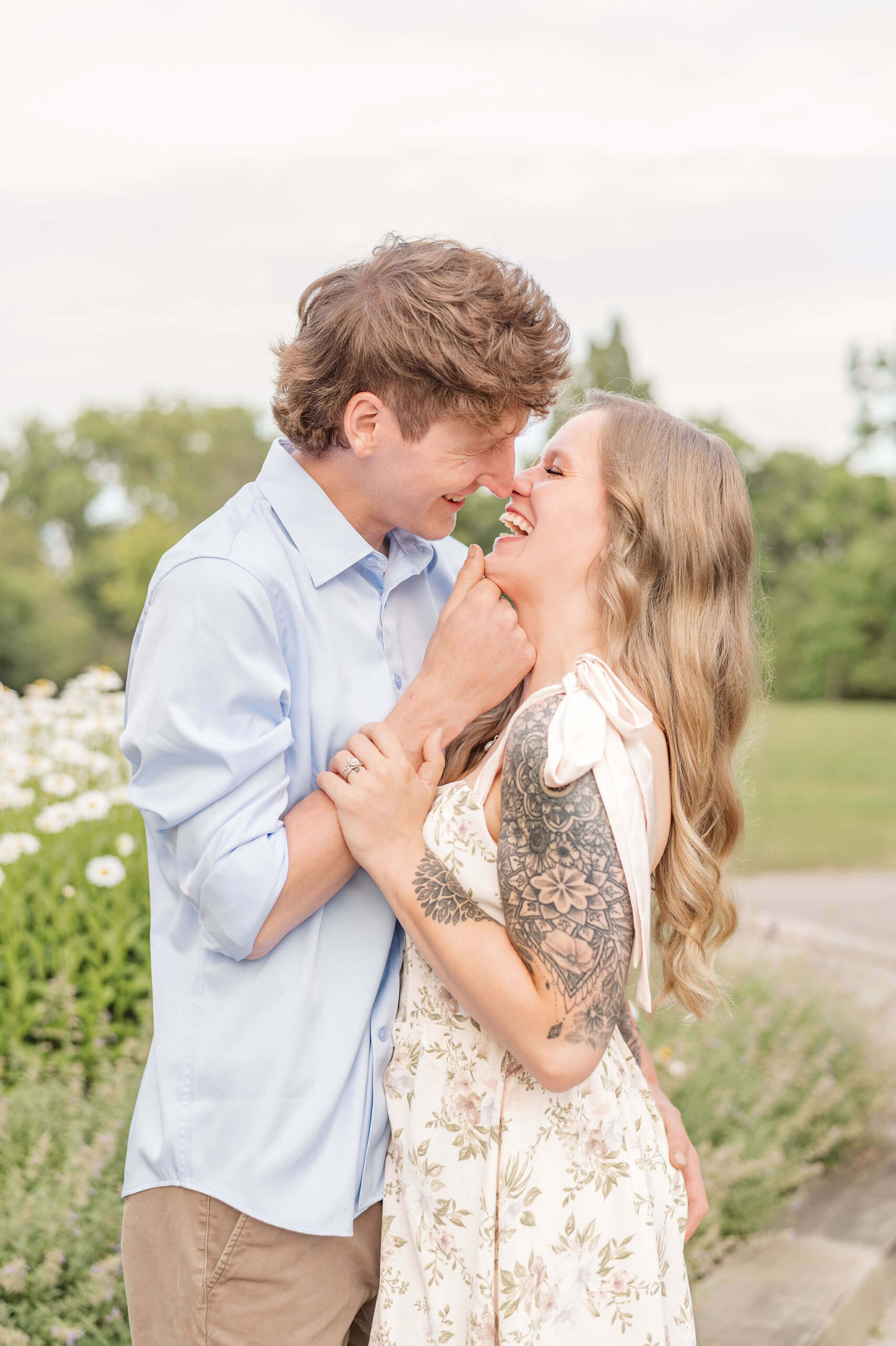 Happy newly engaged couple laugh while leaning in for a kiss during their Pittsburgh engagement photos in a garden in a white dress and blue shirt