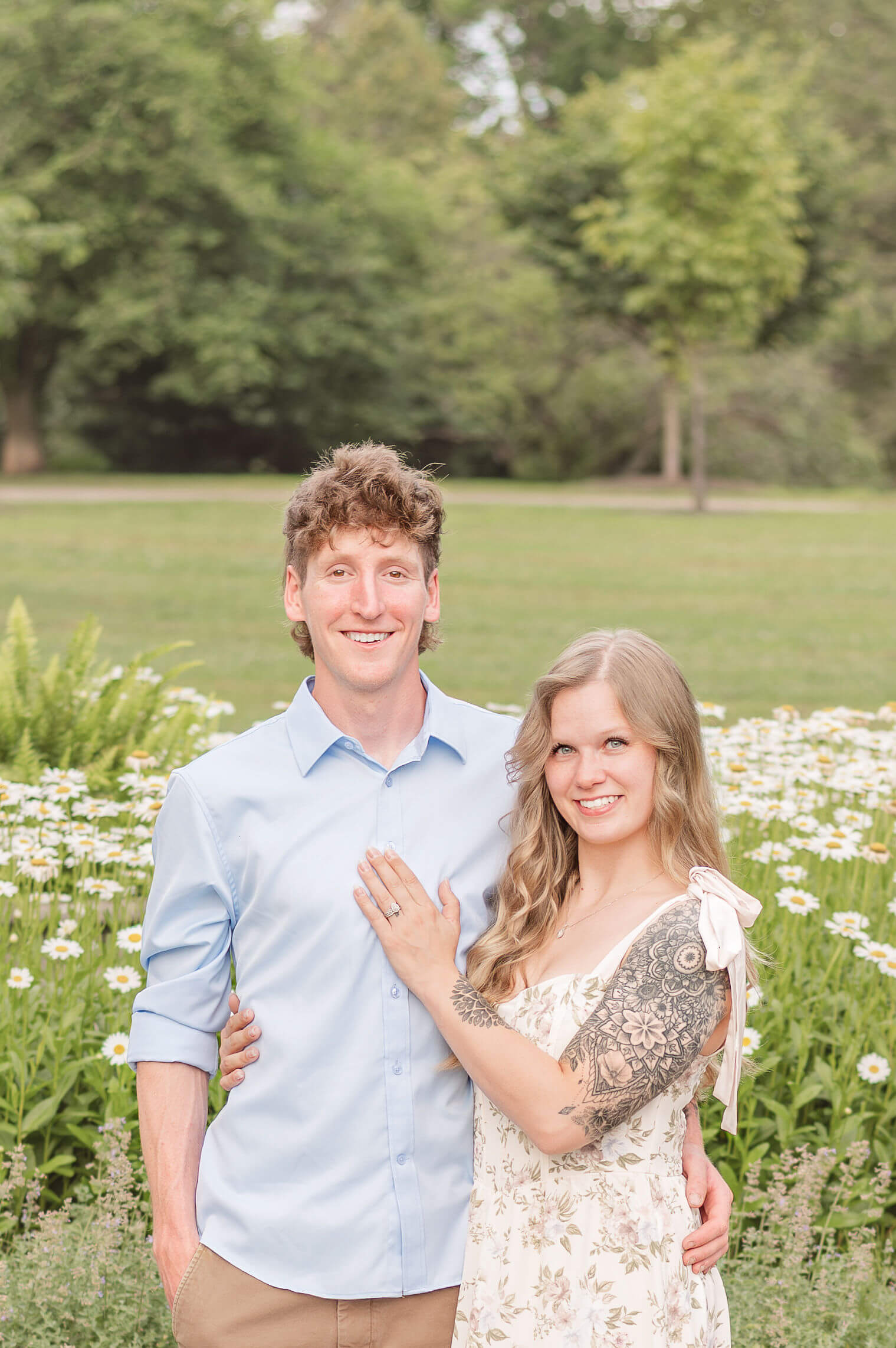 Engaged couple posing in front of daisies, smiling directly at the camera. 