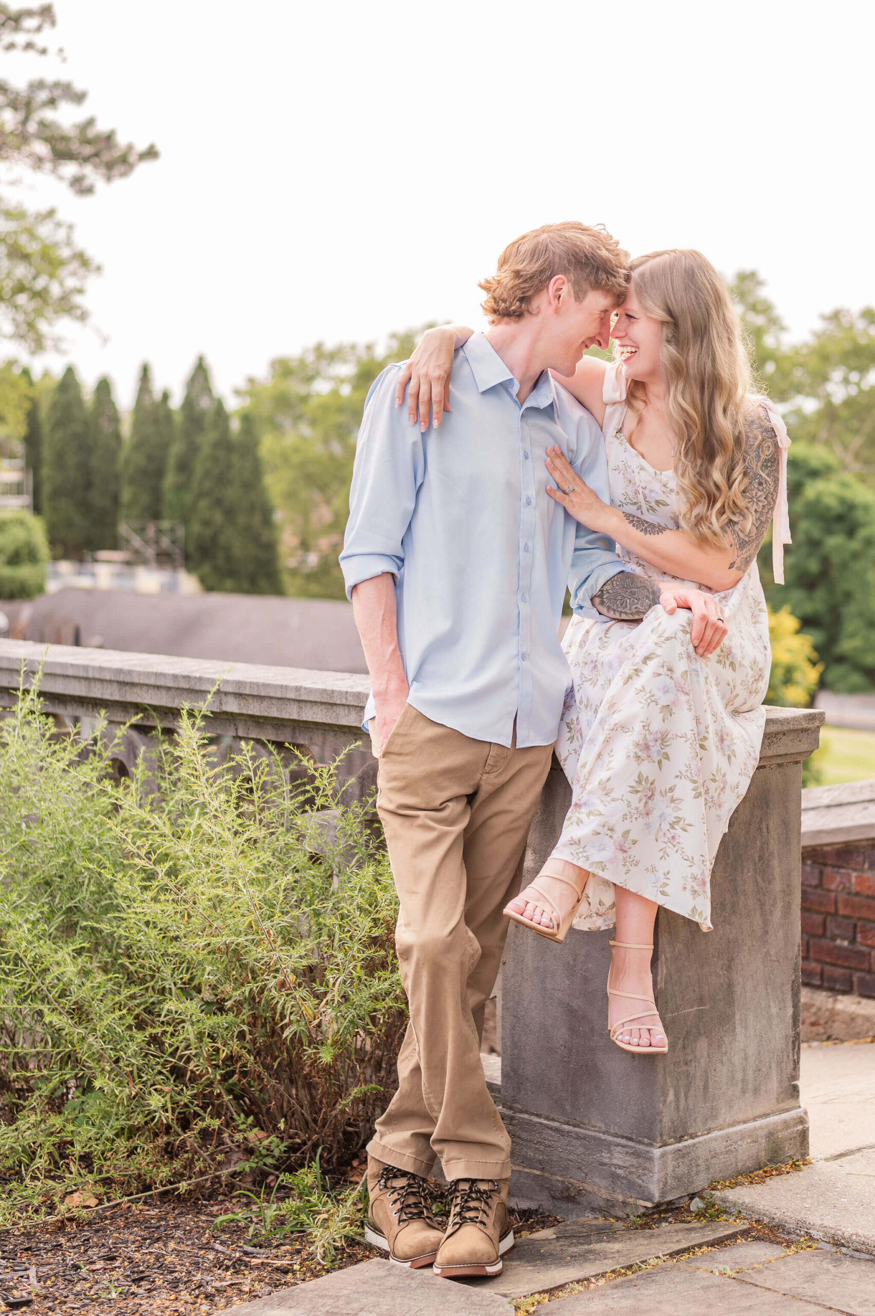 Engaged couple laughing together with bride seated on stone pillar.