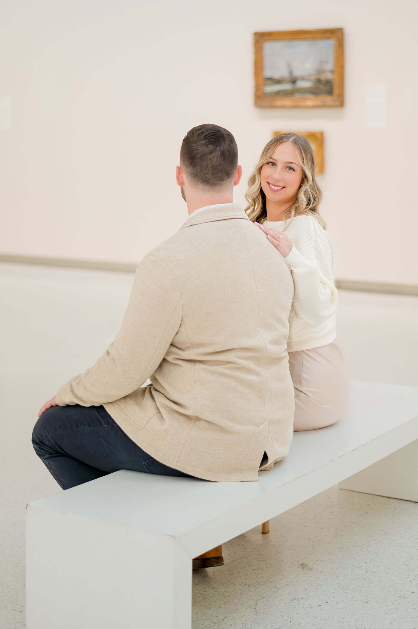 Engaged couple sitting on a white bench inside the Carnegie Museum of Art in Pittsburgh, Pennsylvania, with the bride-to-be smiling at the camera during their engagement session.