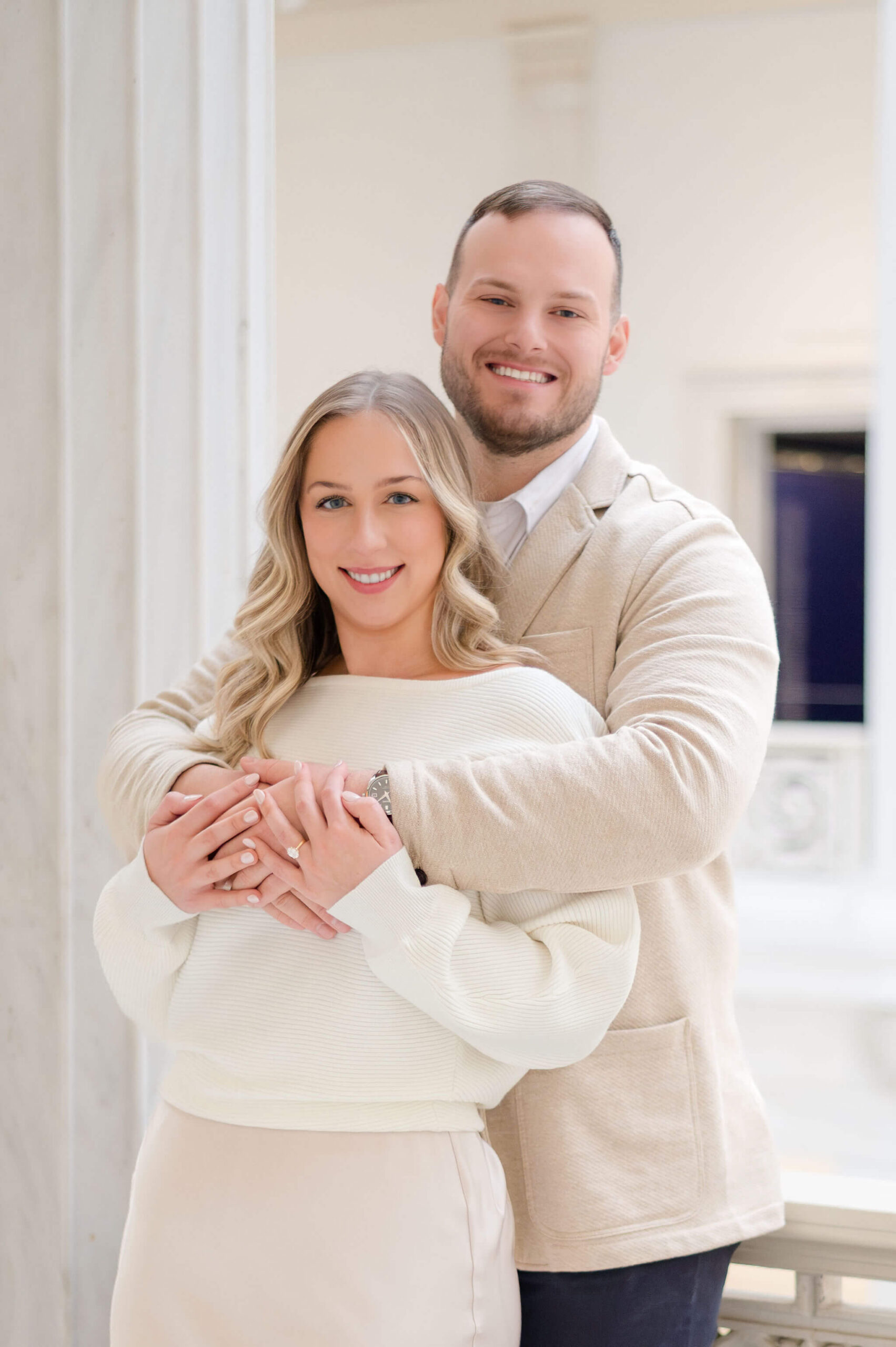 Engaged couple smiling at the camera while embracing inside the Carnegie Museum of Art in Pittsburgh, Pennsylvania, with marble columns in the background.