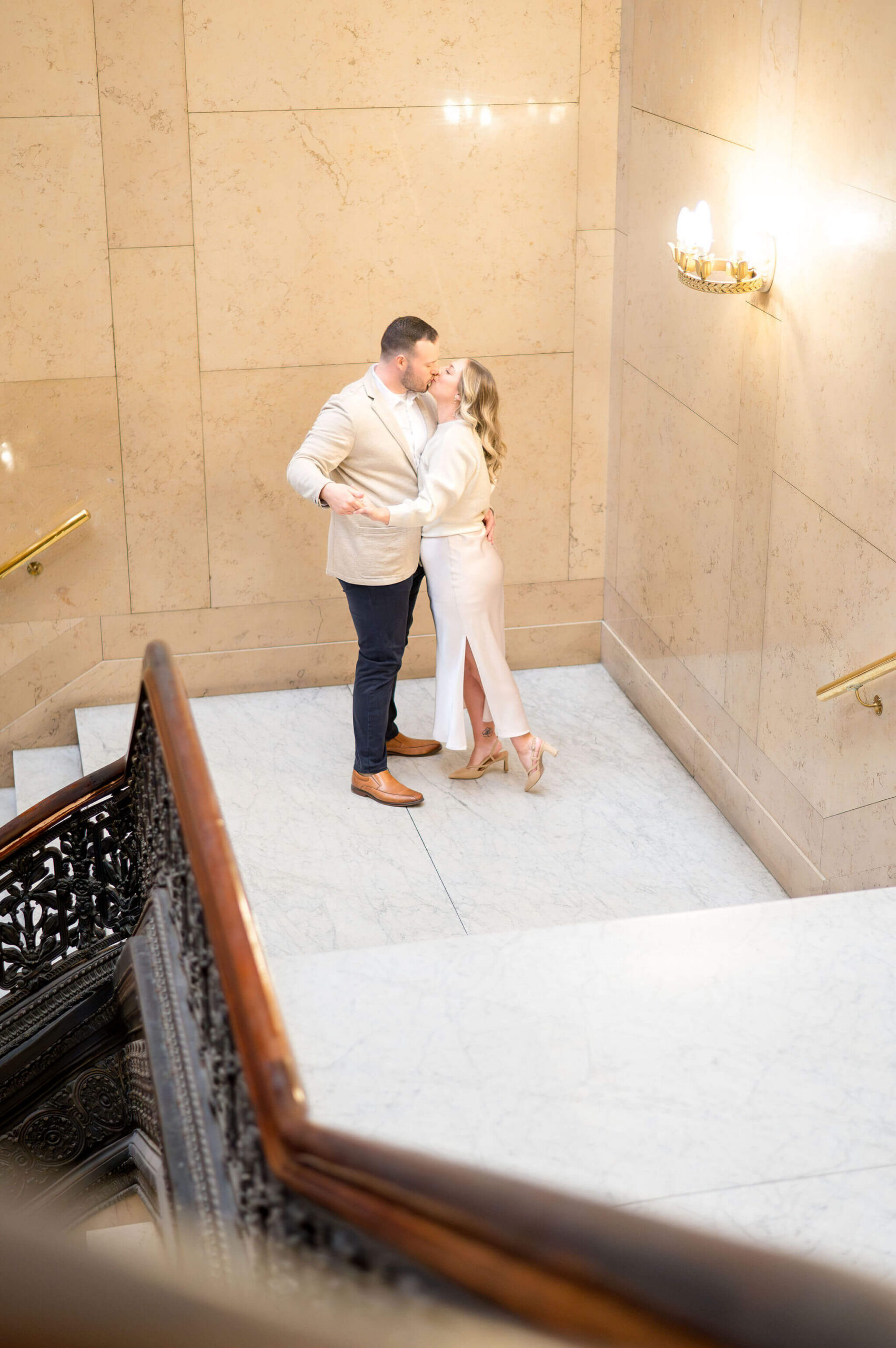 Engaged couple sharing a kiss on a marble staircase landing surrounded by warm stone walls and classic architectural details.