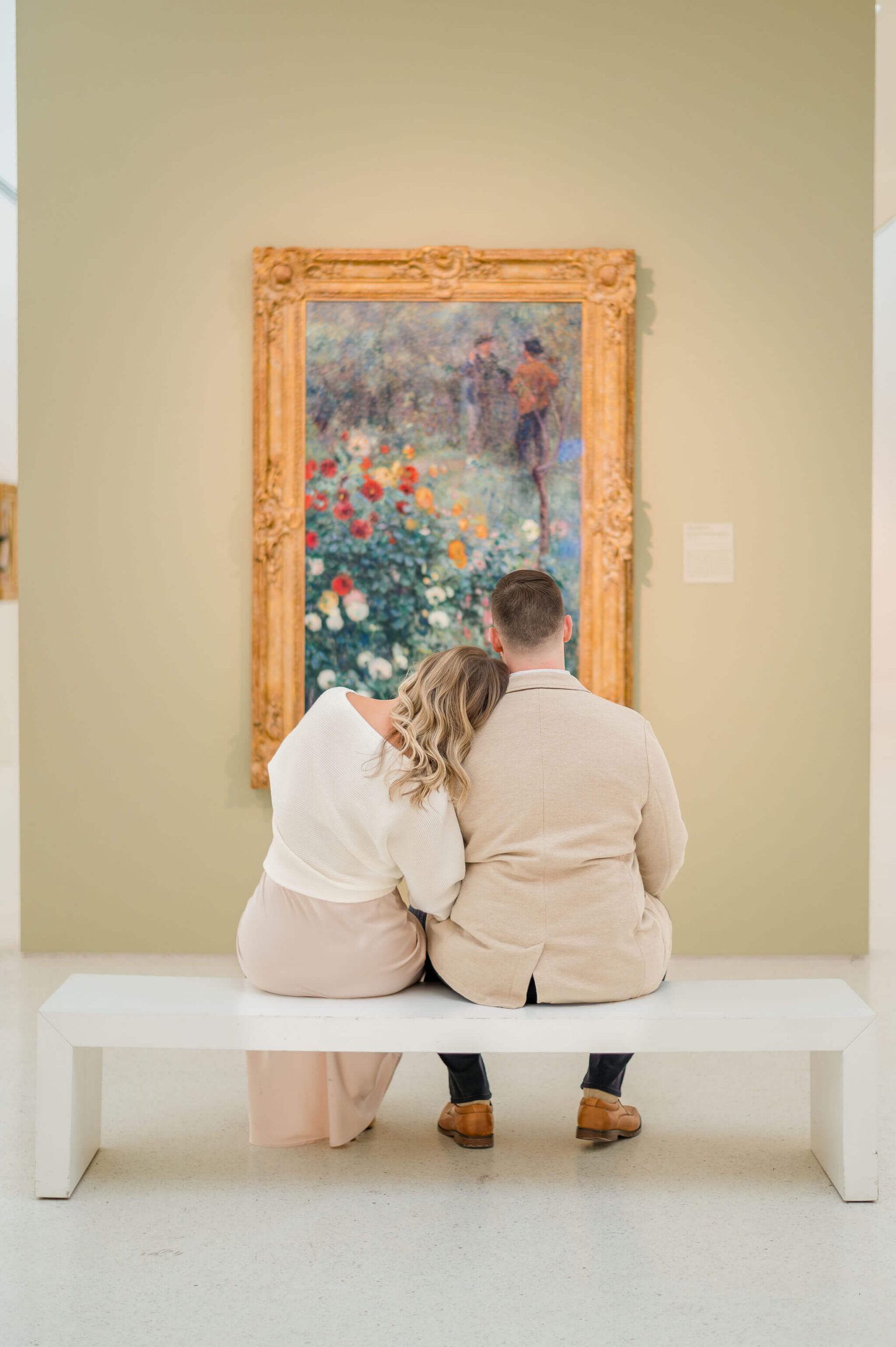 Couple sitting on a white bench inside the Carnegie Museum of Art in Pittsburgh, Pennsylvania, leaning into each other while viewing a framed impressionist painting during their engagement session.