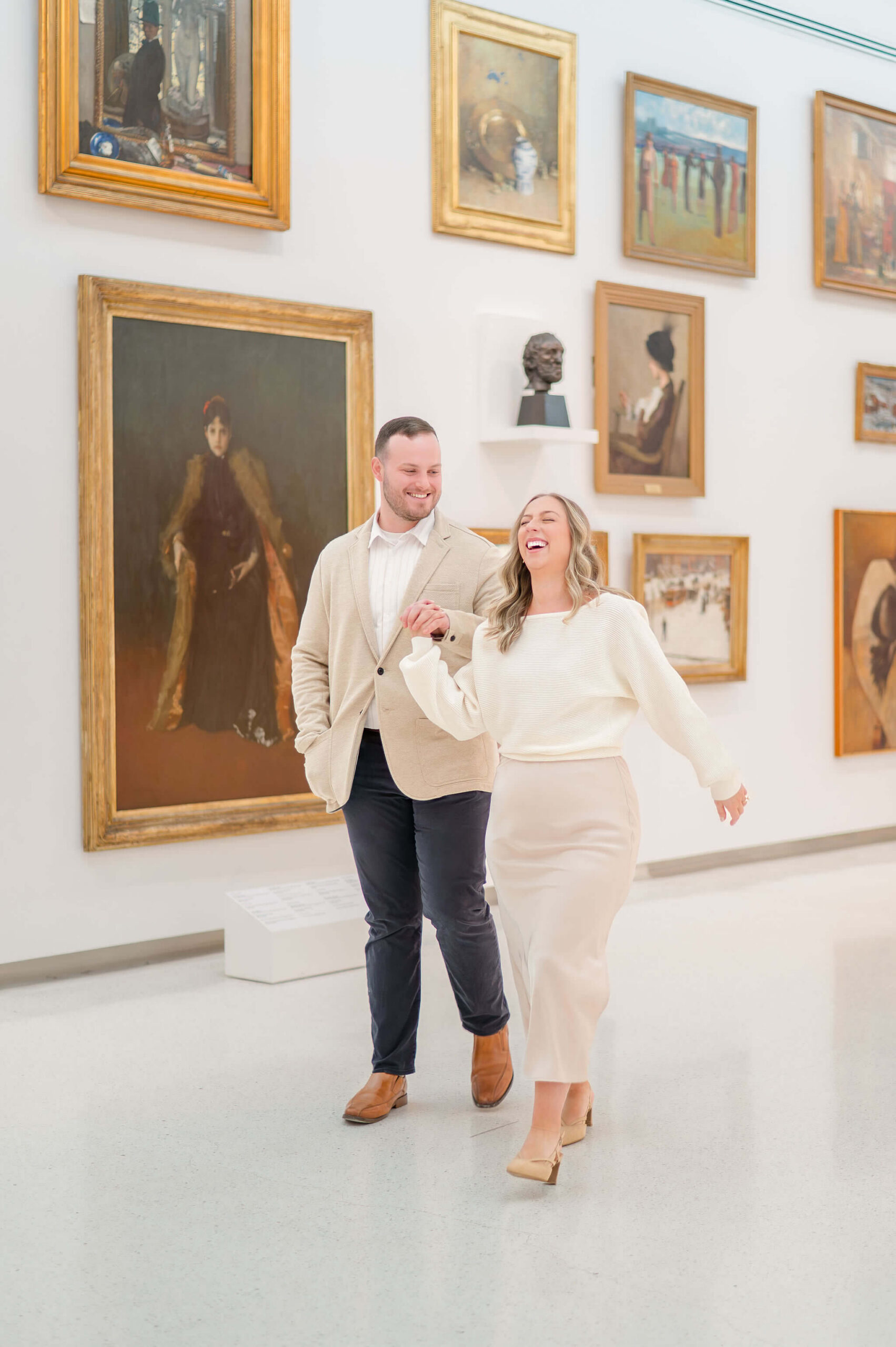 Couple walking hand in hand through an art museum laughing together in front of a gallery wall of framed paintings during their engagement session.